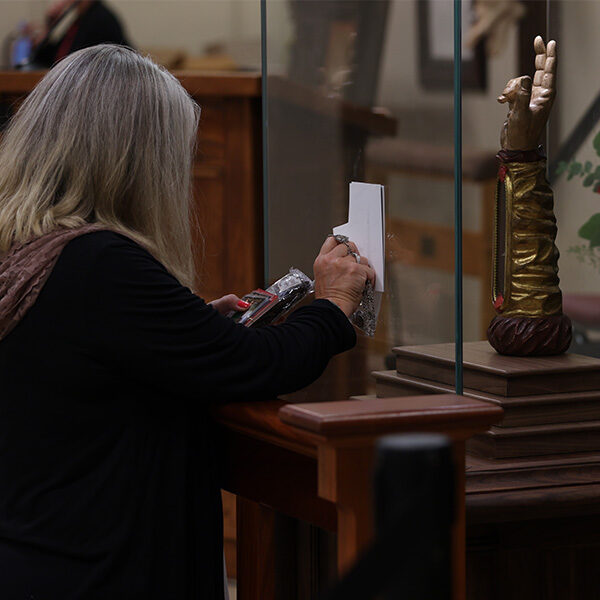 An older woman kneeling to venerate a relic while holding items up to the relic arm of Saint Jude. An older woman kneeling to venerate a relic while holding items up to the relic arm of Saint Jude.