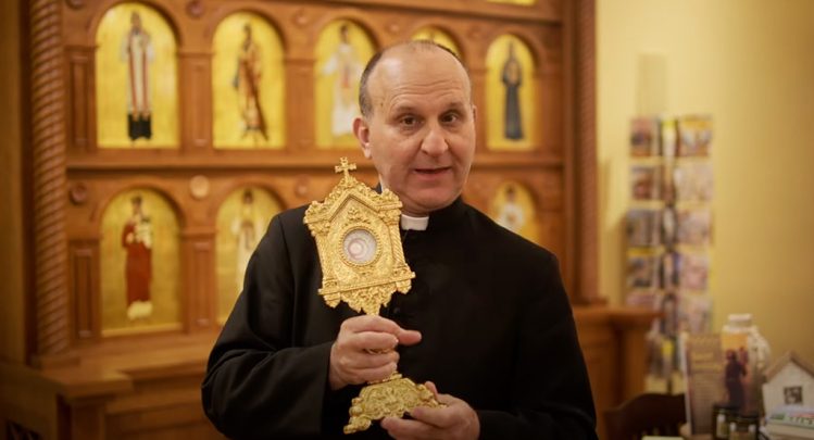 Father Paul Check holding a golden reliquary in a room adorned with sacred artwork, featured in a video titled 'Blessed Carlo Acutis: Living the Christian Life' at the Shrine of Our Lady of Guadalupe in La Crosse, Wisconsin.