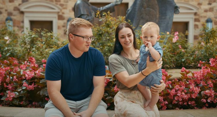 A family sitting in a garden with vibrant flowers at the Shrine of Our Lady of Guadalupe in La Crosse, Wisconsin, featured in a video titled 'Finding the Answer to Prayer'.